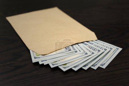 An open brown envelope, crafted from recycled paper, lies atop a stack of US hundred-dollar bills arrayed on a dark wooden surface, highlighting the juxtaposition of sustainable materials and wealth.