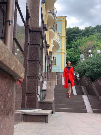 A person in a bright red outfit ascends outdoor stairs beside a building with yellow walls and white balconies. The sky is overcast, and green trees are visible in the background.