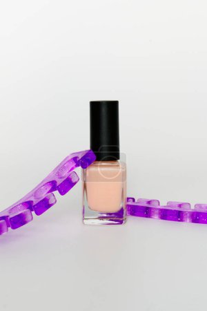 Eine Flasche Pfirsich-Nagellack ist von einer spiralförmigen lila Kette auf weißem Hintergrund umgeben, die den Kontrast und die Lebendigkeit der Farben betont.