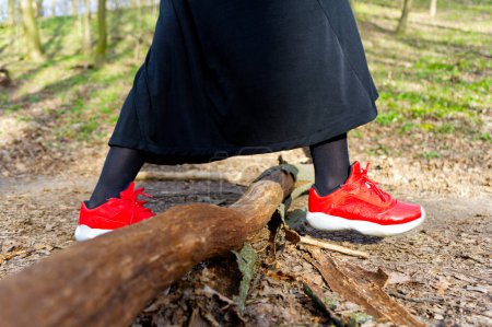 Eine Person balanciert auf einem Baumstamm in einem sattgrünen Wald und trägt leuchtend rote Turnschuhe, die im Kontrast zur natürlichen Umgebung stehen. Die lebendigen Schuhe verleihen dieser ruhigen Waldlandschaft eine dynamische Note