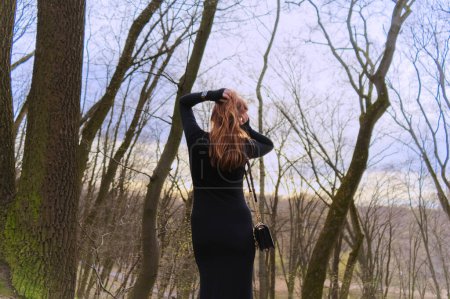 A person with not long hair, dressed in black, stands amidst bare trees, gazing into a forested area. The overcast sky and leafless branches create a serene yet somber atmosphere.