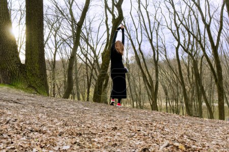 A person in a black dress stretches an arm upwards amidst bare trees in a forest, capturing a moment of tranquility and connection with nature.
