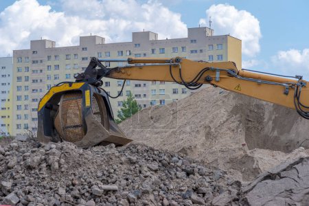 Road building. Heaps of stones and substrate. Visible excavator arm with rotating screen. Road construction work in the city among blocks of flats. Under a blue sky with soft white fluffy clouds. Excavator arm with soil fractionation equipment.