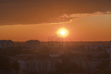 Juni-Abend, extrem farbenfroher - in Gold- und Orangetönen - Sonnenuntergang über der Stadt (Ostrowiec). Ein bewölkter Himmel, der von der untergehenden Sonne vergoldet wird, eine Stadt, die langsam in der Dunkelheit des Abends unten versinkt.