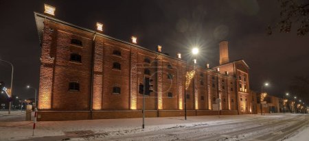 Old historic restored brewery illuminated at night by tiny spotlights. City in the snow.A dark night sky with clouds swollen with snow. Below you can see the historic brick brewery building and freshly fallen snow all around. 