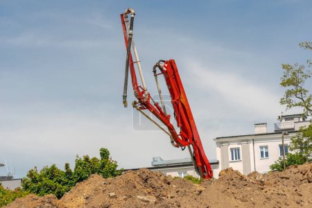 . Excavator working on a construction site.Red concrete pump on a road construction site.Pouring concrete on a road construction site - a red concrete pump is straightening its pipes, against the background of a blue, slightly cloudy sky . 