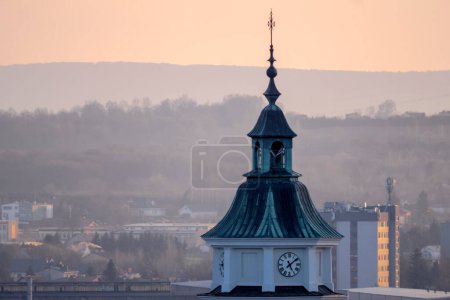The tower of the city of the old town.The tower with a clock of a historic church on a foggy early spring day. A church tower with a roof covered with copper sheets, hills and a mountain range in the background.  