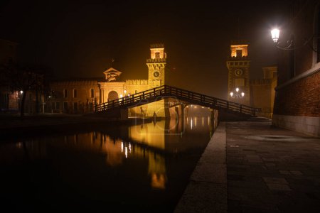This is an image of the Arsenale, Venice, Italy. The image was taken from the front and was shot at night when the structure and surrounding area was illuminated. It includes the wooden bridge spanning the canal with reflections on its surface.