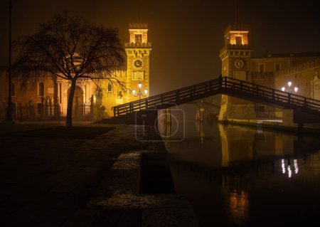 This is an image of the Arsenale, Venice, Italy. The image was taken from the front and was shot at night when the structure and surrounding area was illuminated. It includes the wooden bridge spanning the canal with reflections on its surface.