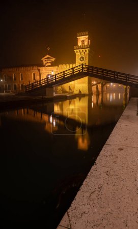 Esta es una imagen del Arsenale, Venecia, Italia. La imagen fue tomada desde el frente y fue filmada por la noche cuando la estructura y el área circundante fueron iluminados. Incluye el puente de madera que atraviesa el canal con reflejos en su superficie.