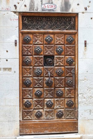 This is an image of a highly decorative wood door.. The main section is divided into squares. Every other square has an embossed metal disc in the same design. In the center is a peephole constructed of metal and designed to look like an eye.