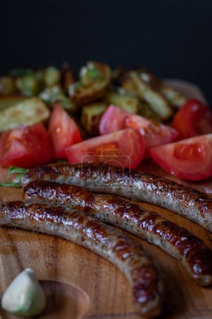 Grilled sausages served with roasted potato wedges, fresh tomato slices, and a clove of garlic on a wooden cutting board.