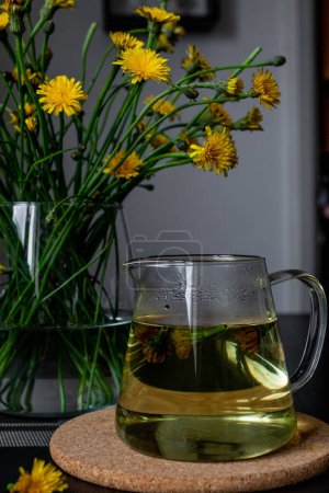 Une cruche en verre remplie de thé de pissenlit est assise sur une table, avec des fleurs de pissenlit fraîches en arrière-plan. Le thé pissenlit est connu pour ses bienfaits pour la santé, y compris ses propriétés diurétiques et détoxifiantes.