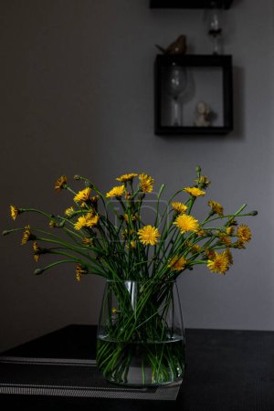 A glass jug filled with dandelion tea sits on a table, with fresh dandelion flowers in the background. Dandelion tea is known for its health benefits, including its diuretic and detoxifying properties.