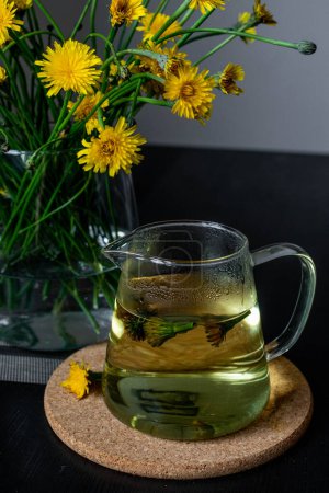 A glass jug filled with dandelion tea sits on a table, with fresh dandelion flowers in the background. Dandelion tea is known for its health benefits, including its diuretic and detoxifying properties.