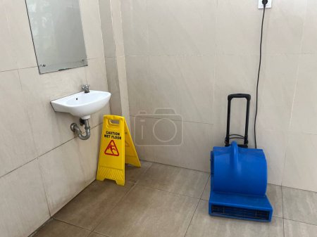 Vorsicht nasser Fußboden gelbe Warntafel unter dem Waschbecken mit blauem Fußbodentrockner in öffentlicher Toilette