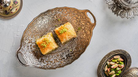 Foto de Trozos de dulces orientales con pistachos en un plato blanco sobre un suelo blanco rodeado de utensilios de plata y cobre - Imagen libre de derechos