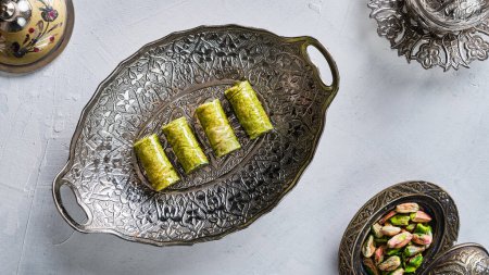 Foto de Trozos de dulces orientales con pistachos en un plato blanco sobre un suelo blanco rodeado de utensilios de plata y cobre - Imagen libre de derechos