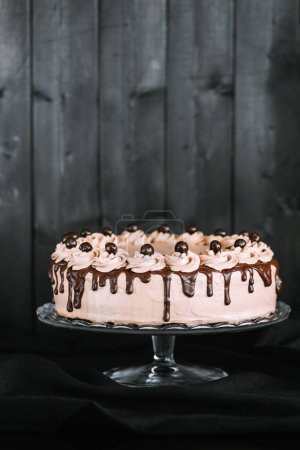 A chocolate cake with chocolate icing on a glass cake stand against dark wooden background.