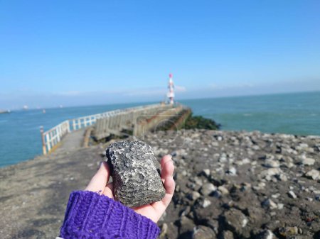 A woman's hand holding a geocache that looks like a black stone which was hidden between rocks on a beach on a sunny day in Netherlands