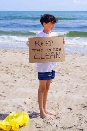 boy picking up plastic on the beach and holding up a sign saying KEEP THE BEACH CLEAN
