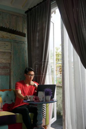 Photo for Asian Man Working on Laptop in Sunlit Villa, Sofa Comfort, Productivity - Royalty Free Image