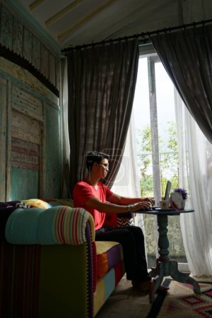 Photo for Asian Man Working on Laptop in Sunlit Villa, Sofa Comfort, Productivity - Royalty Free Image