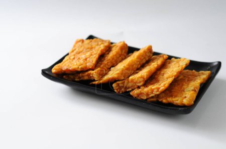 Foto de Tempeh frito servido en un plato negro, fondo blanco aislado, concepto tradicional de alimentos - Imagen libre de derechos