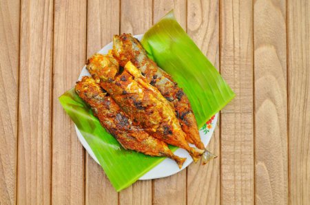 Ikan Bakar oder Gegrillter Fisch mit indonesischen Gewürzen auf Bananenblatt und isoliertem Holzhintergrund.