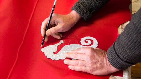 Foto de Un sastre traza una plantilla de un caballito de mar sobre un lienzo rojo con su mano y un lápiz - Imagen libre de derechos