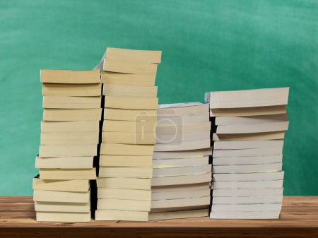 A pile of books laying on a wooden table with a green board in the background.