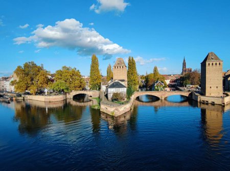 Ponts Couverts de Strasbourg. Eine steinerne Brücke mit drei Türmen überspannt einen blauen Fluss. Saftig grüne Bäume säumen das Flussufer auf beiden Seiten. Im Hintergrund thront eine steinerne Burg mit einem hohen zentralen Turm auf einem Hügel..