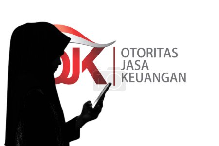 Foto de Indonesia-13 de marzo de 2024: una mujer que sostiene un teléfono inteligente frente a una pantalla grande con el logotipo de otoritas jasa keuangan - Imagen libre de derechos