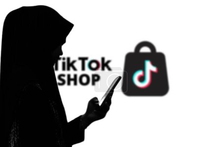 Téléchargez les photos : Une femme tenant un smartphone devant un grand écran avec le logo de TikTokshop en arrière-plan. Indonésie-13 mars 2024. - en image libre de droit