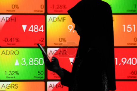 Téléchargez les photos : Indonésie - 28 février 2024 : Femme musulmane tenant un smartphone devant un grand écran avec la Bourse d'Indonésie - en image libre de droit