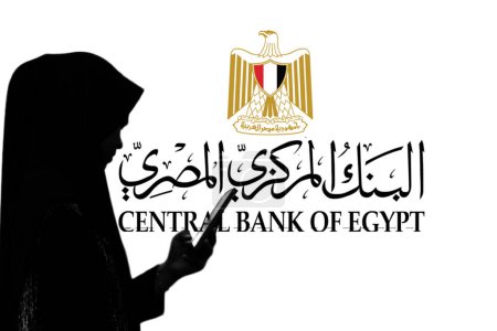 Téléchargez les photos : Indonésie - 4 avril 2024 : Femme tient un smartphone devant un grand écran avec le logo de la Banque centrale d "Égypte en arrière-plan. - en image libre de droit