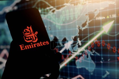Foto de Indonesia - 7 de abril de 2024: teléfono inteligente de mano Con el logotipo de Emirates Airlines se muestra en la pantalla del teléfono inteligente Con el mercado de valores en segundo plano. - Imagen libre de derechos