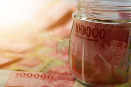 Photo for Indonesia - April 7th 2024: The concept of saving for finance accounting involves placing Indonesian banknotes in the denomination of 100,000 rupiah into a glass jar. - Royalty Free Image