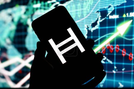 Foto de Indonesia-25 de abril 2024: el logotipo de Hedera, HBAR, criptomoneda hedera, logotipo de criptomoneda se muestra en la pantalla del teléfono inteligente con el mercado de valores en el fondo. - Imagen libre de derechos