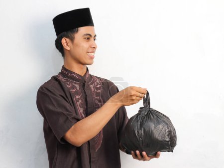 Portrait of Asian Muslim man holding and giving a black bag, depicted with a friendly and smiling expression, isolated on a white background.Kurban and zakat Ramadan concept