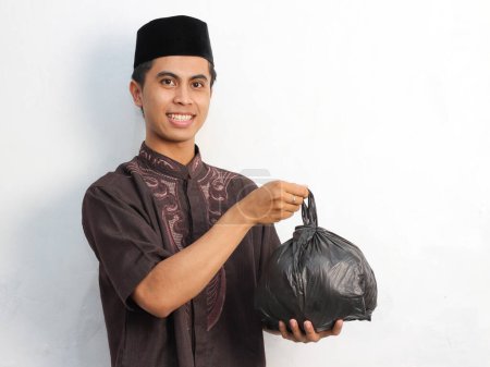 Portrait d'un homme musulman asiatique tenant et donnant un sac noir, représenté avec une expression amicale et souriante, isolé sur un fond blanc. Concept Kurban et zakat Ramadan