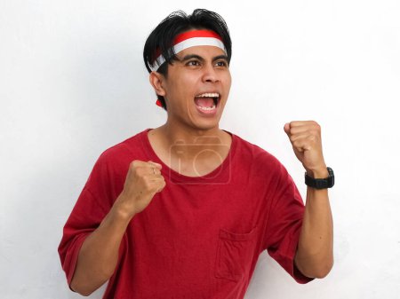 Téléchargez les photos : Portrait d'un jeune homme asiatique portant un t-shirt rouge frappant et un bandeau avec le drapeau indonésien. Levant les deux poings avec enthousiasme, célébrant l'indépendance de l'Indonésie 17 août. - en image libre de droit