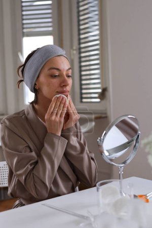 Téléchargez les photos : Femme peau nettoyante avec de l'eau micellaire et coton Pad et regarde dans le miroir. Photo de haute qualité - en image libre de droit