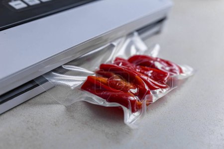 Téléchargez les photos : Machine à sceller emballage sous vide avec sac en plastique de poivron rouge à table blanche, gros plan. Photo de haute qualité - en image libre de droit
