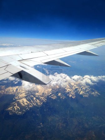 Téléchargez les photos : Vue de la fenêtre de l'avion aile de l'avion et des montagnes avec des sommets enneigés blancs au-dessous du ciel bleu avion de passagers - en image libre de droit