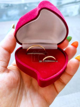 Foto de La chica tiene en su mano una caja roja con dos anillos de compromiso para las propuestas de boda preparativos de boda mañana de la novia, caja de terciopelo en forma de corazón rojo - Imagen libre de derechos