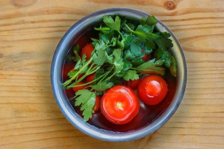 Fresh vegetables are being washed  in the metal bowl filled with cold water on the vintage cracked wooden cutting board. Organic tomatoes, parsley, celery. Top view. Copy space