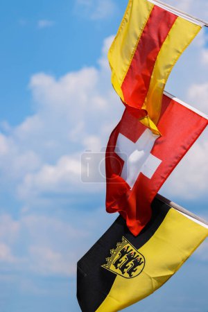 Three flags on the flagpoles - waving in the wind. Flags of Baden, Switzerland, Flag Baden-Wrttemberg with an emblem. Swiss flag in the middle. Isolated on the blue sky and white clouds. Copy space.