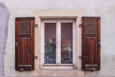 Fenster im Landhausstil mit alten getrockneten Blumen, hölzernen Fensterläden im Retro-Stil, einem alten Foto hinter Glas und weißen Netzvorhängen (Tüll). Altdeutsches historisches Haus in Bayern. Shabby strukturierte Wand.
