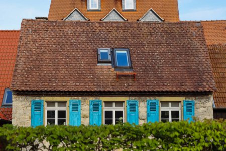 Fassade des alten gelben Fachwerkhauses mit gelber Steinmauer, rotem Ziegeldach und blauen Fensterläden. Grüne Büsche vor dem Haus. Sommertag in Ochsenfurt, Bayern.
